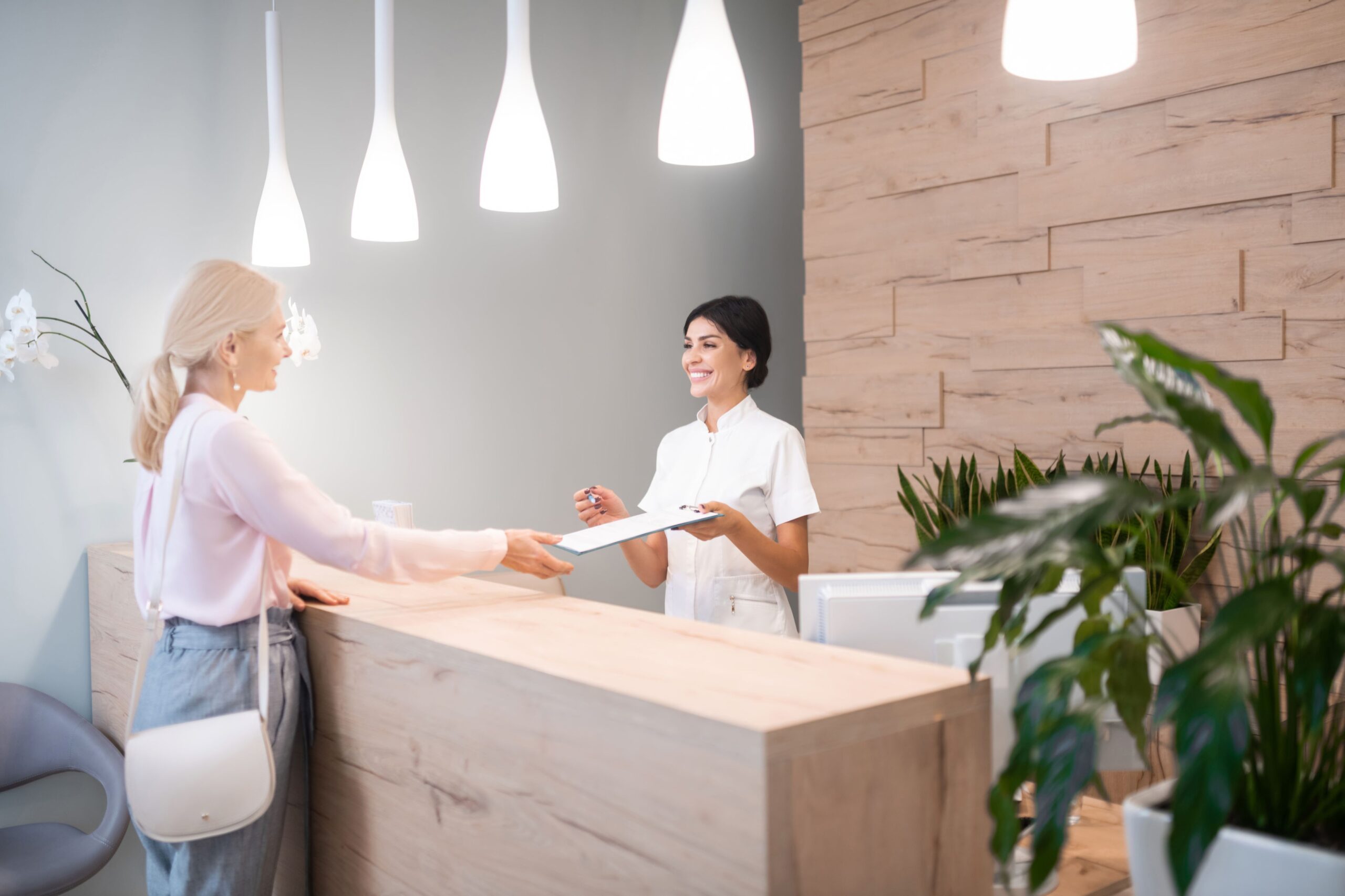 woman-standing-at-reception-of-dental-clinic-2023-11-27-05-25-03-utc Receptionist in white handing a document to a woman in pink at a modern reception desk. The environment is bright, welcoming, and professional.