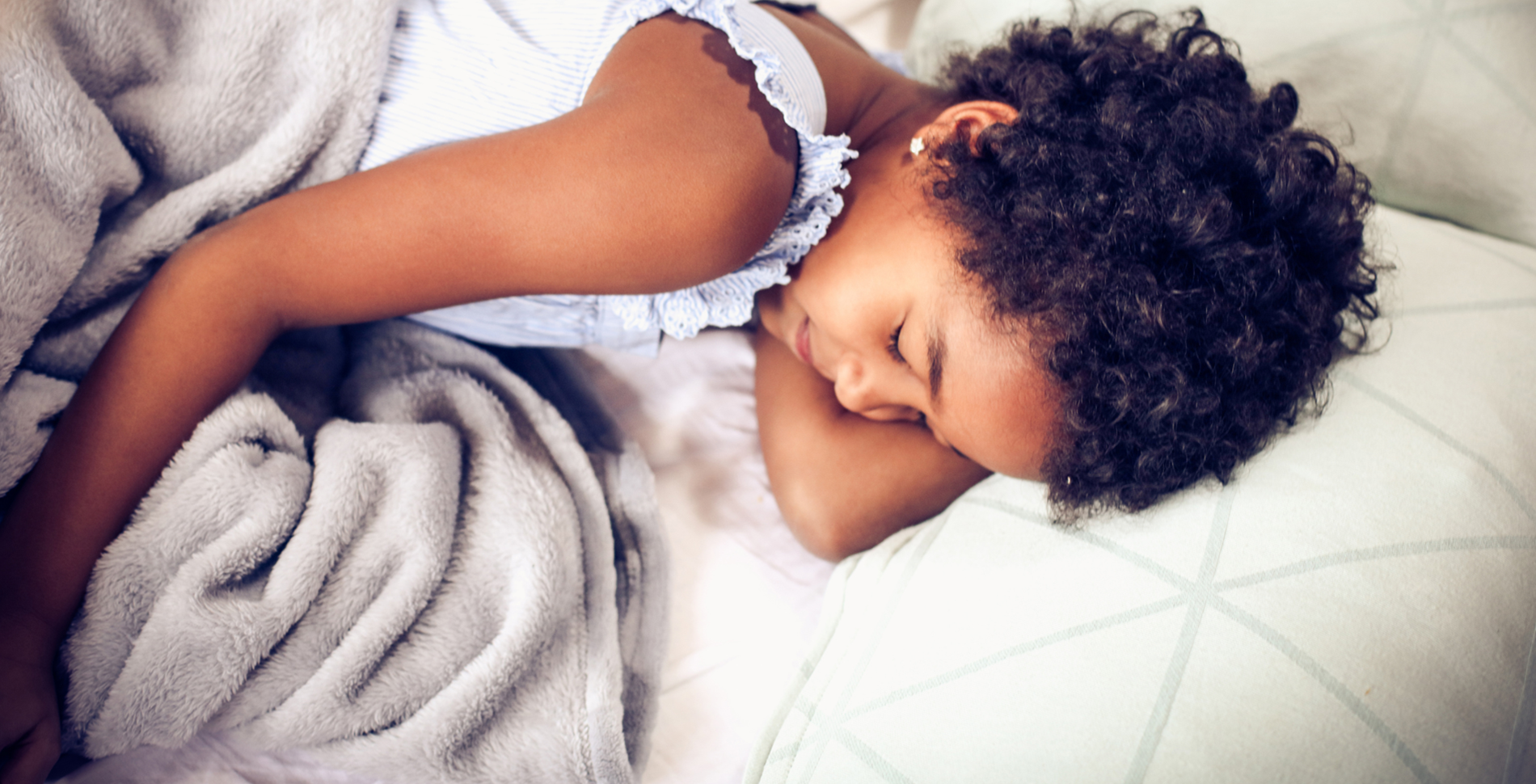 A child sleeps peacefully, nestled in a soft gray blanket on a bed with white, patterned sheets. The scene conveys warmth and serenity.