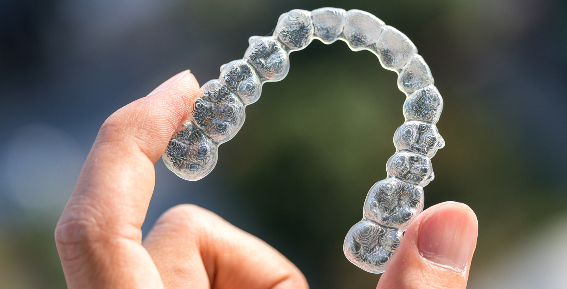 Clear Aligners Invisalign-blog-img A hand holds a clear, curved dental aligner against a blurred outdoor background. The transparent device subtly reflects light, conveying precision.