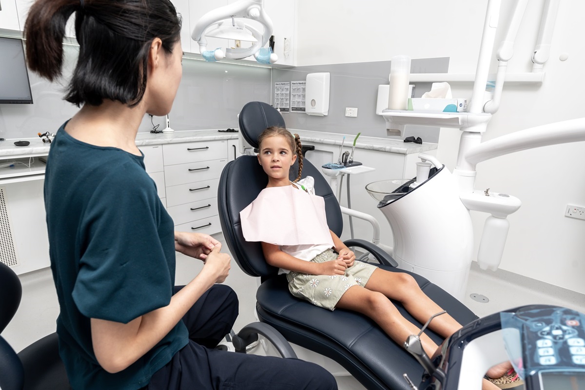 A young girl sits in a dental chair, wearing a pink bib, while a female dentist in scrubs talks to her, creating a calm and reassuring atmosphere. A young girl sits in a dental chair, wearing a pink bib, while a female dentist in scrubs talks to her, creating a calm and reassuring atmosphere.