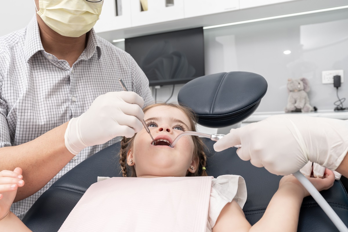 A child sits in a dental chair with a pink bib, while a dentist examines her teeth using dental tools. The setting is a bright, clean dental office. A child sits in a dental chair with a pink bib, while a dentist examines her teeth using dental tools. The setting is a bright, clean dental office.