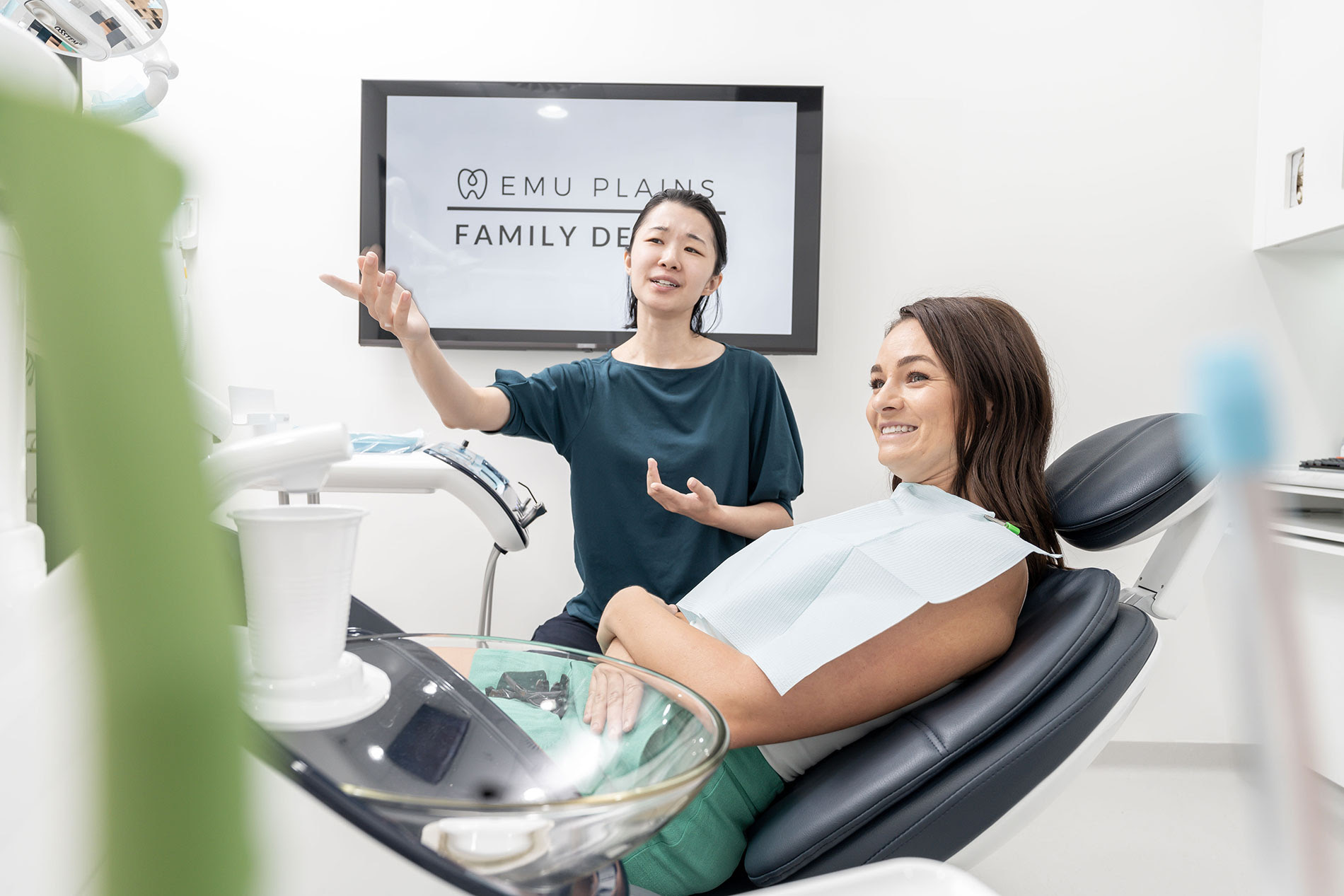 A dentist gestures while explaining to a cheerful patient in a dental chair. The clinic has a clean, modern look. Sign reads "Emu Plains Family Dentist."
