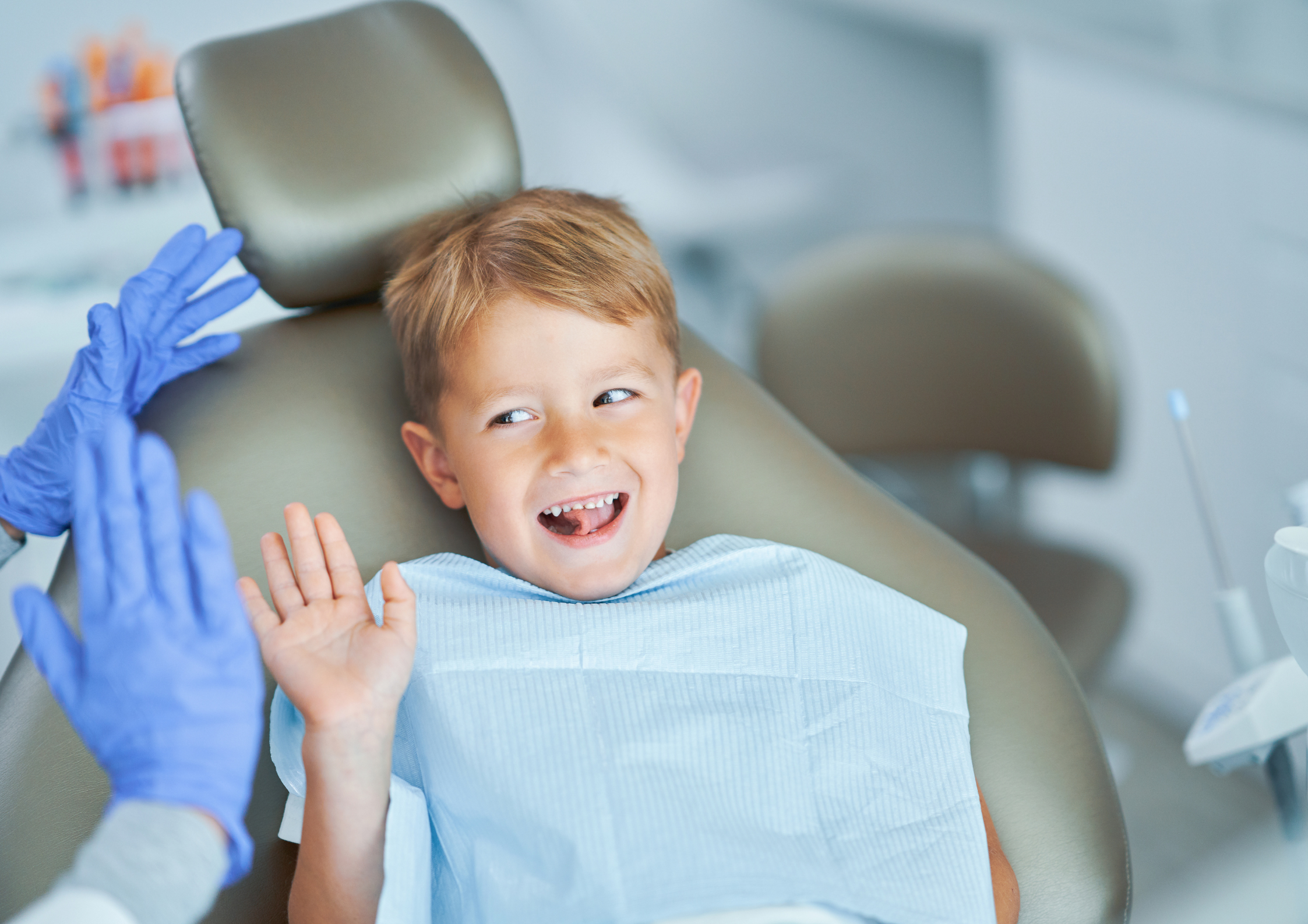 Young boy in a dental chair smiling nervously, wearing a blue bib. Blue-gloved dentist's hands are shown, creating a warm, humorous atmosphere.