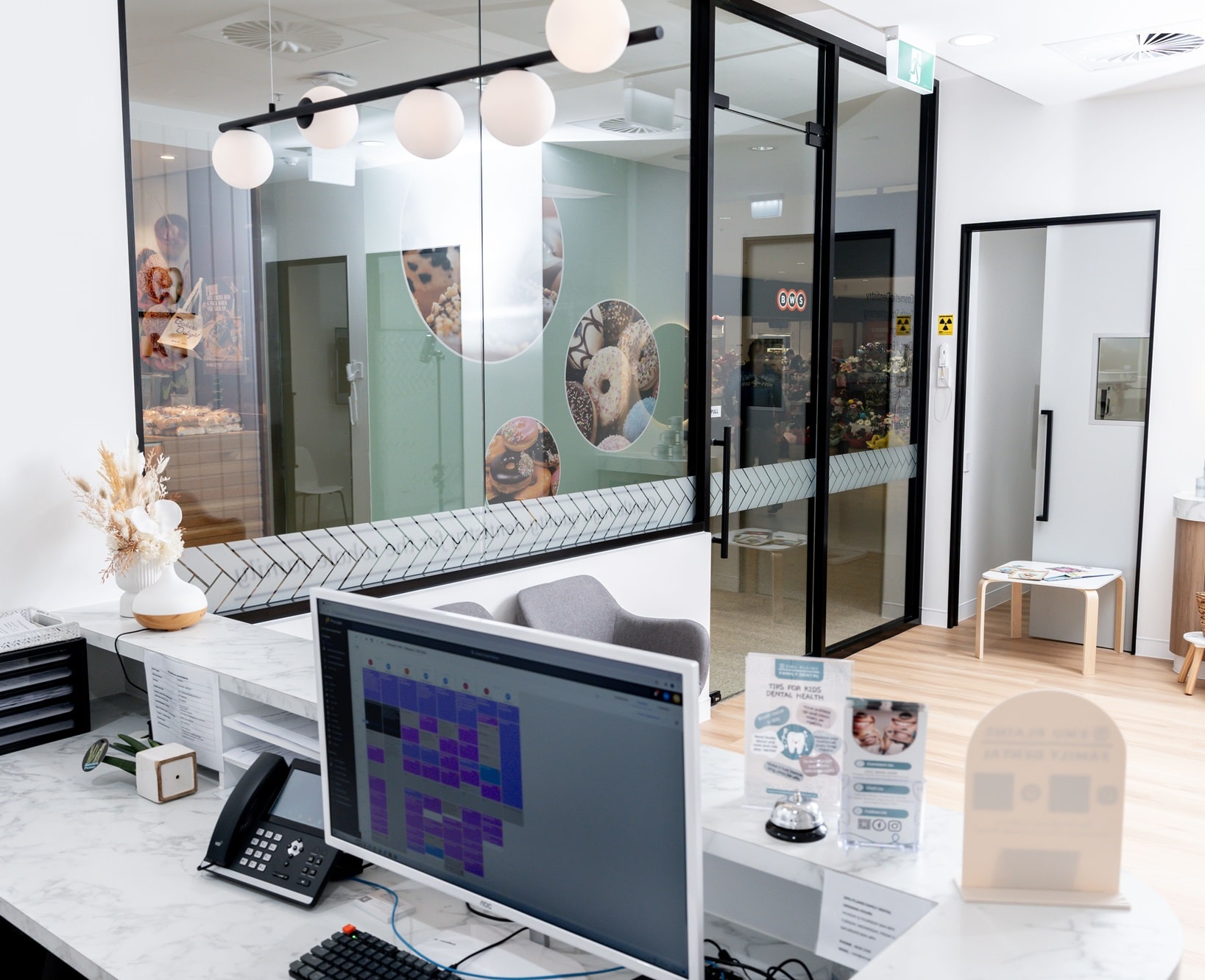 A bright, modern office reception area features a white front desk with a computer, phone, and brochures. Glass walls display playful donut graphics.