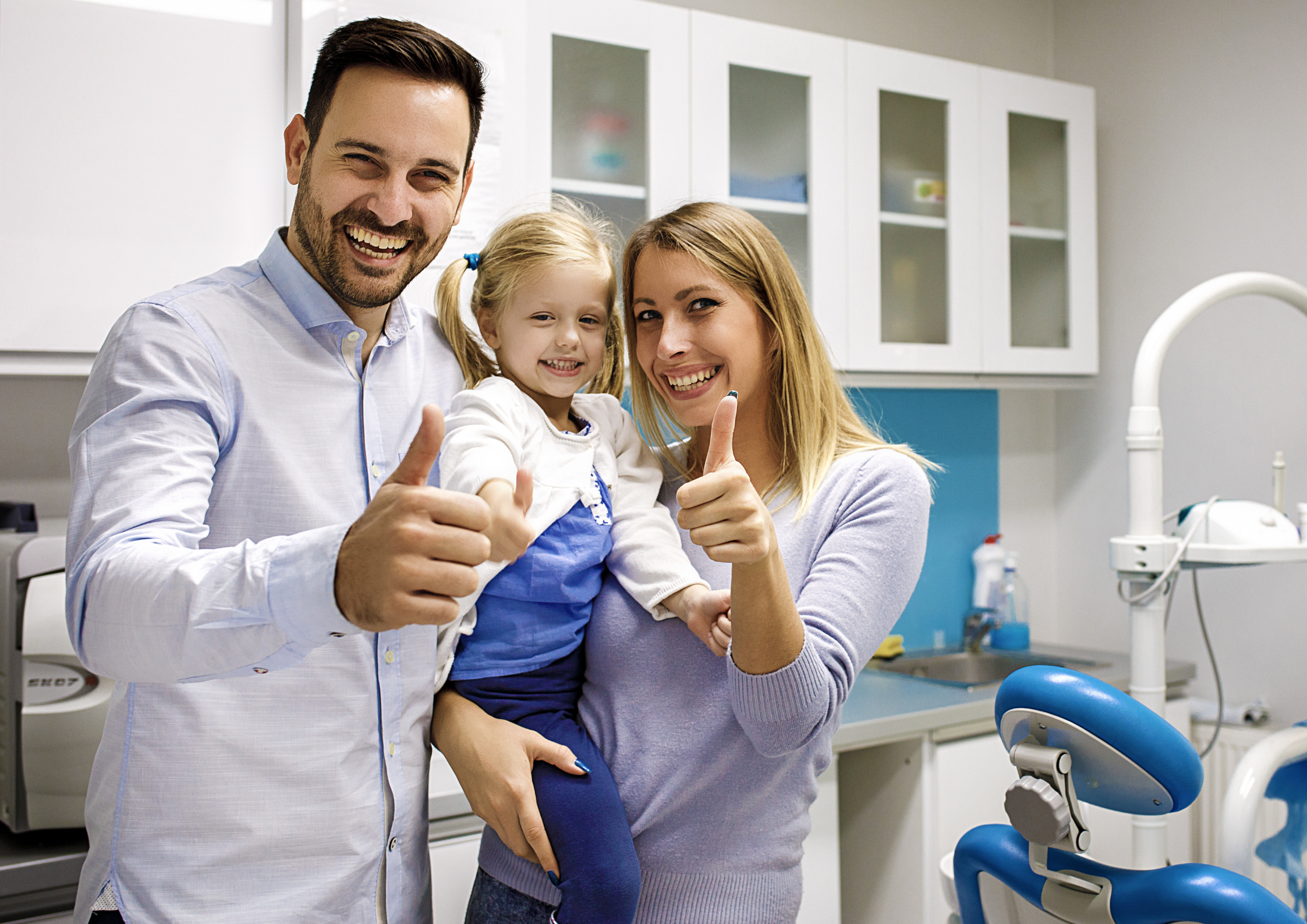 A smiling man and woman give a thumbs-up while holding a young girl in a dental office. The atmosphere is cheerful and supportive. A smiling man and woman give a thumbs-up while holding a young girl in a dental office. The atmosphere is cheerful and supportive.