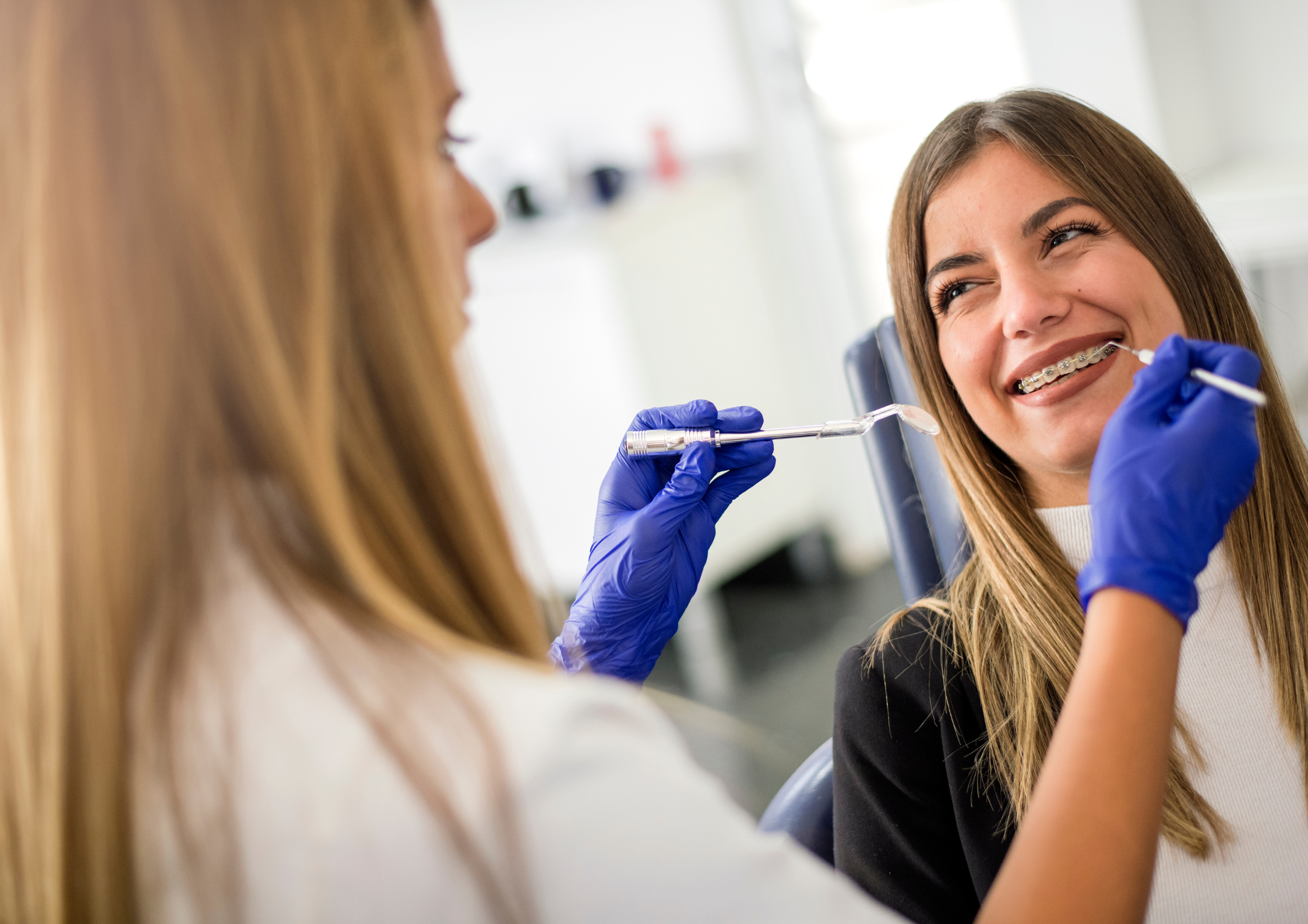 A smiling woman with braces sits in a dental chair as a dentist in blue gloves uses tools. The scene conveys a relaxed, positive atmosphere.