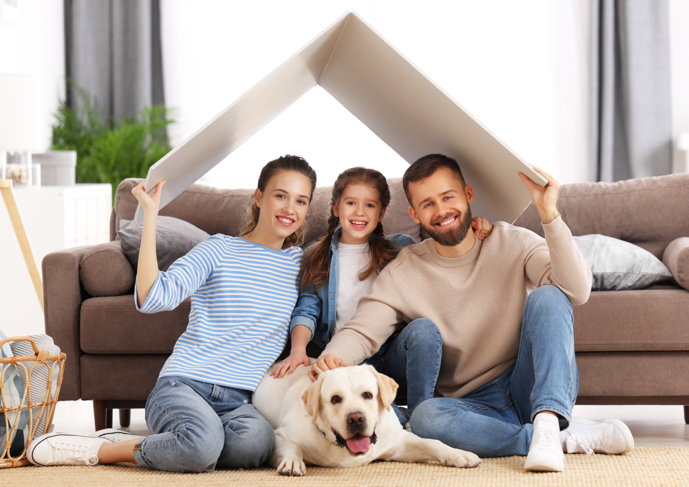A smiling family of three and a dog sit on the floor under a cardboard roof. The setting is a cozy living room, conveying warmth and togetherness. A smiling family of three and a dog sit on the floor under a cardboard roof. The setting is a cozy living room, conveying warmth and togetherness.