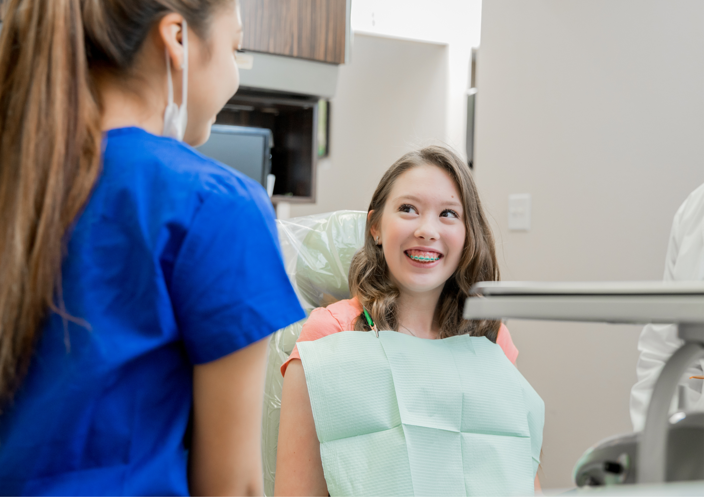 A girl with braces smiles at a dental professional in blue scrubs, conveying a friendly and reassuring atmosphere in a dental office setting.