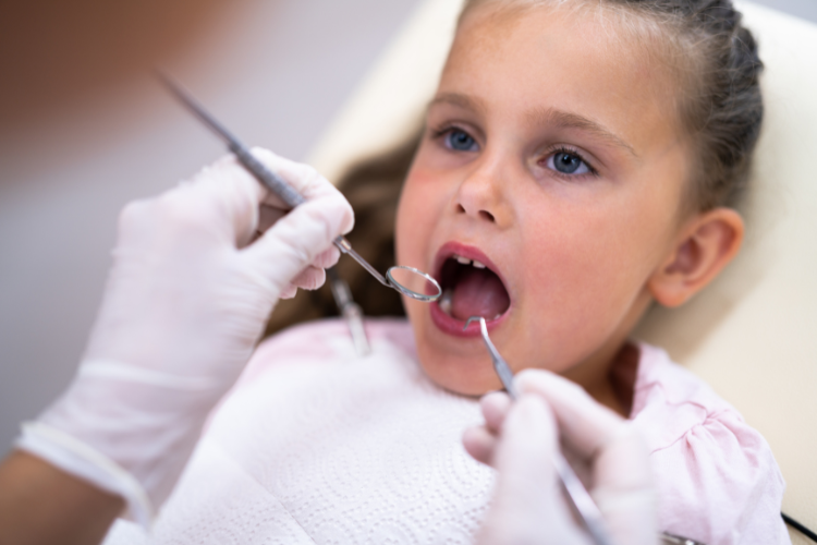 A young girl sits in a dentist's chair, mouth open. A dentist wearing gloves uses dental tools for an examination. The scene feels calm and professional.