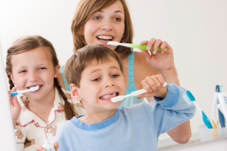 A woman and two children are brushing their teeth, smiling in a bright bathroom. The kids, one girl and one boy, wear pajamas, conveying a cheerful, morning routine.