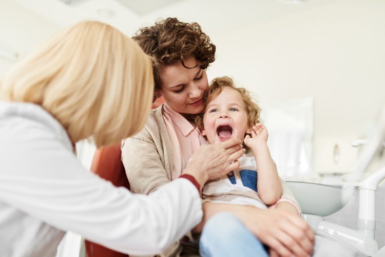 A dentist in a white coat examines a smiling child's teeth. The child sits on a woman's lap, looking content in a bright, friendly dental office.