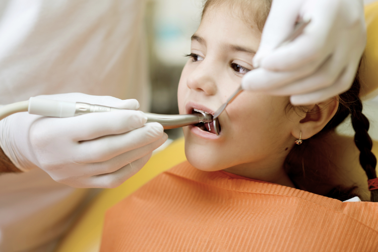 A young girl at the dentist, wearing an orange bib. The dentist, in white gloves, uses dental tools to examine her mouth. The atmosphere is calm and clinical.