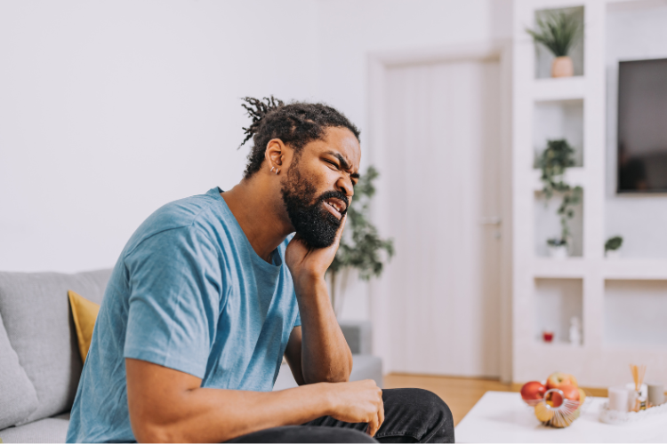 Wisdom Teeth and Dental Health: When to Act and When to Wait Man in a blue t-shirt sitting on a couch, grimacing in pain while holding his neck. The room is bright with plants and shelves in the background.