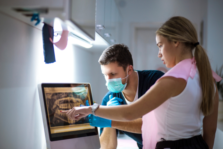 A dentist and a patient examine a dental X-ray on a computer screen. The dentist, in gloves and mask, points to the image, indicating focused attention.