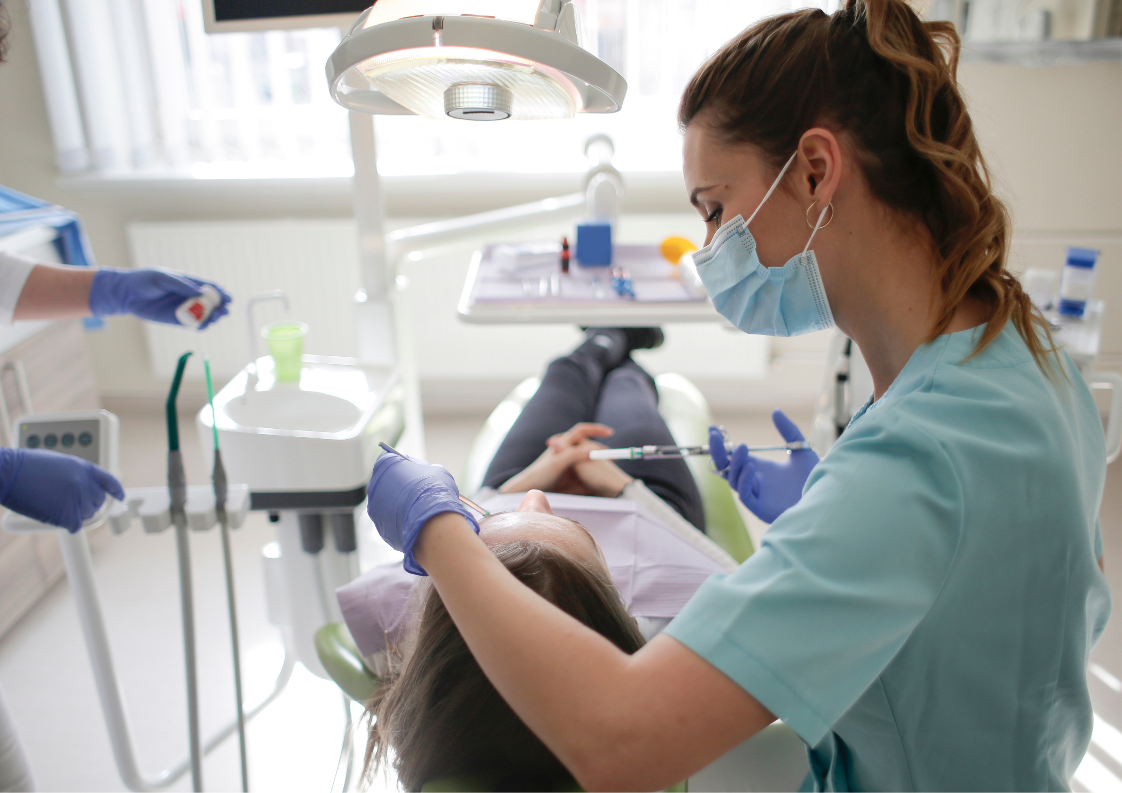 Dentist wearing a mask and gloves prepares to treat a patient lying in a dental chair, with a bright, clinical setting and focused atmosphere.