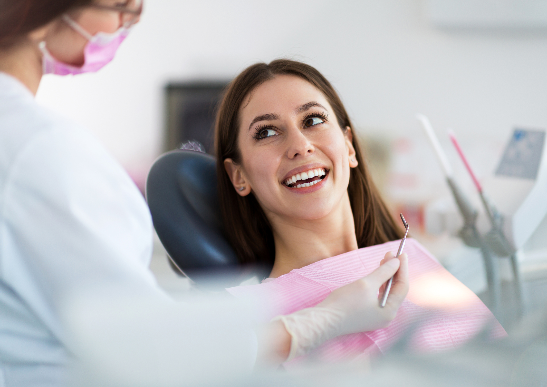 Smiling woman in a dental chair, wearing a pink bib, looks at a dentist holding a tool. The setting is bright and clinical, conveying a relaxed tone.