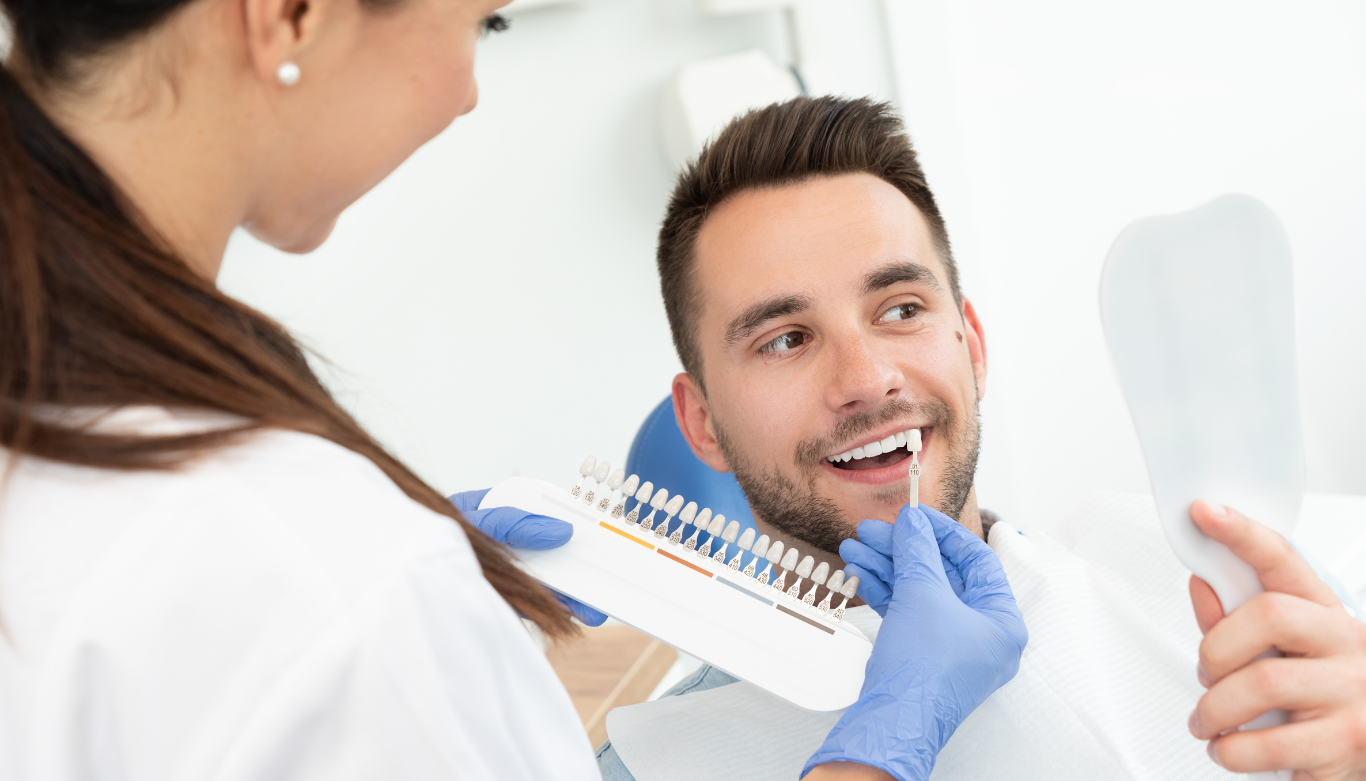 Dental Blog Images 1 A dentist wearing blue gloves holds a teeth shade guide next to a smiling man's mouth. The setting is a dental office, conveying a cheerful tone.