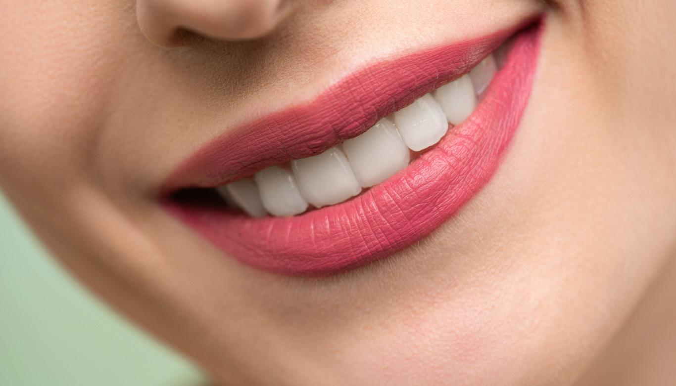 Close-up of a smiling mouth with bright pink lipstick and white teeth. The soft-focus background is light green, conveying a fresh and cheerful tone.