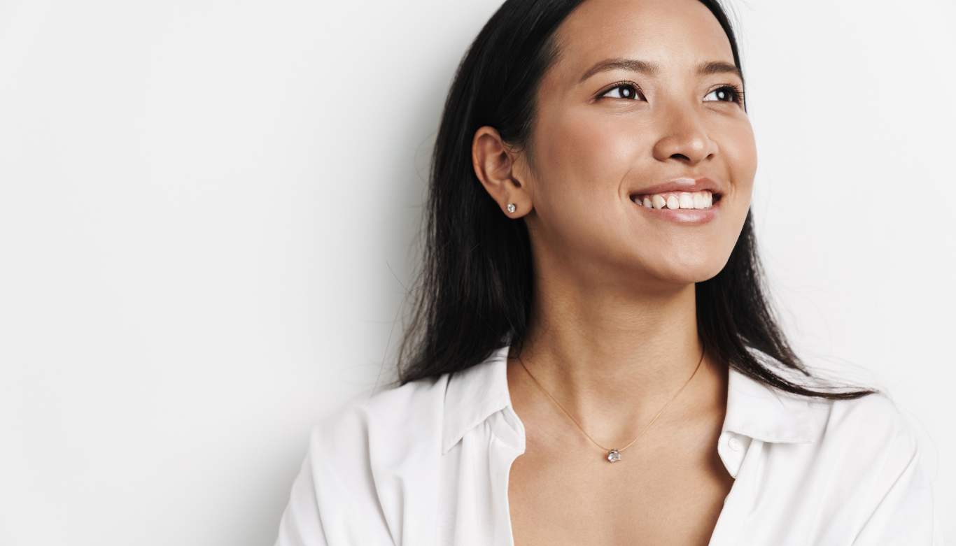 Dental Blog Images 4 Smiling woman looking upward, wearing a white shirt and small earrings against a plain white background, conveying a sense of optimism and contentment.