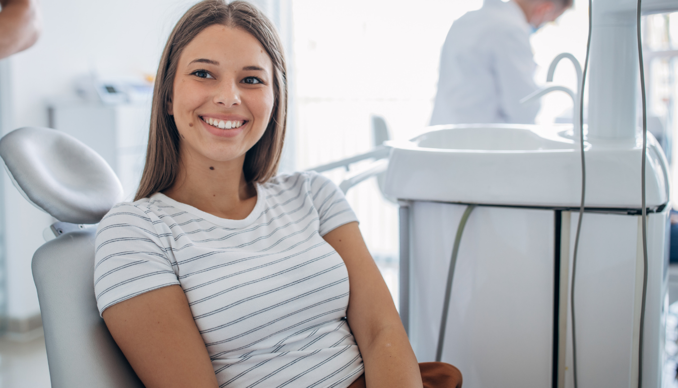 Dental Blog Images 5 A woman with long brown hair smiles while sitting in a dental chair, wearing a white striped shirt. The clinic setting is bright and modern.