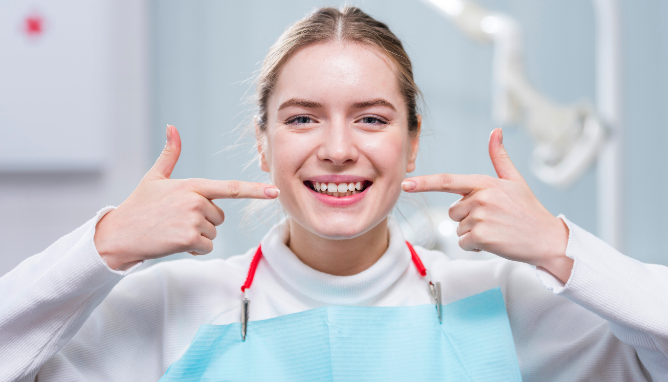 Smiling woman in a dental clinic wearing a blue bib, pointing to her teeth.
